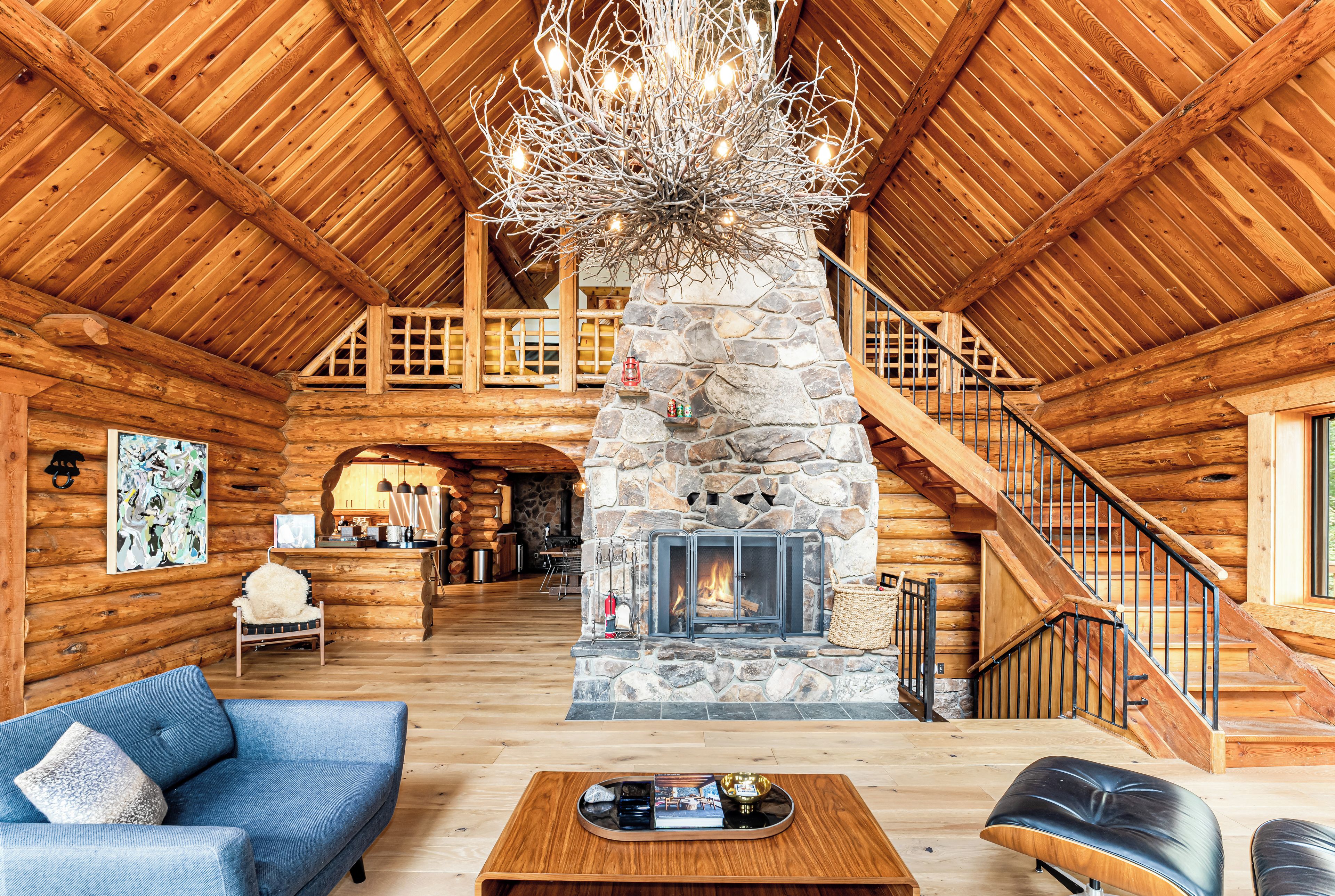 A log cabin interior with a stone fireplace, wooden staircase, and a chandelier.