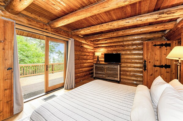 A log cabin bedroom with a bed, a dresser, a television, and a sliding glass door leading to a deck.