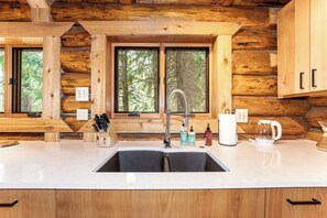Una cocina con un lavamanos doble, armarios de madera y ventana con vista a un bosque.