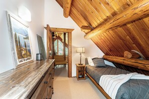 A bedroom with a sloped ceiling, wooden beams, a bed with a quilt, a nightstand, a lamp, a dresser, and a mirror.