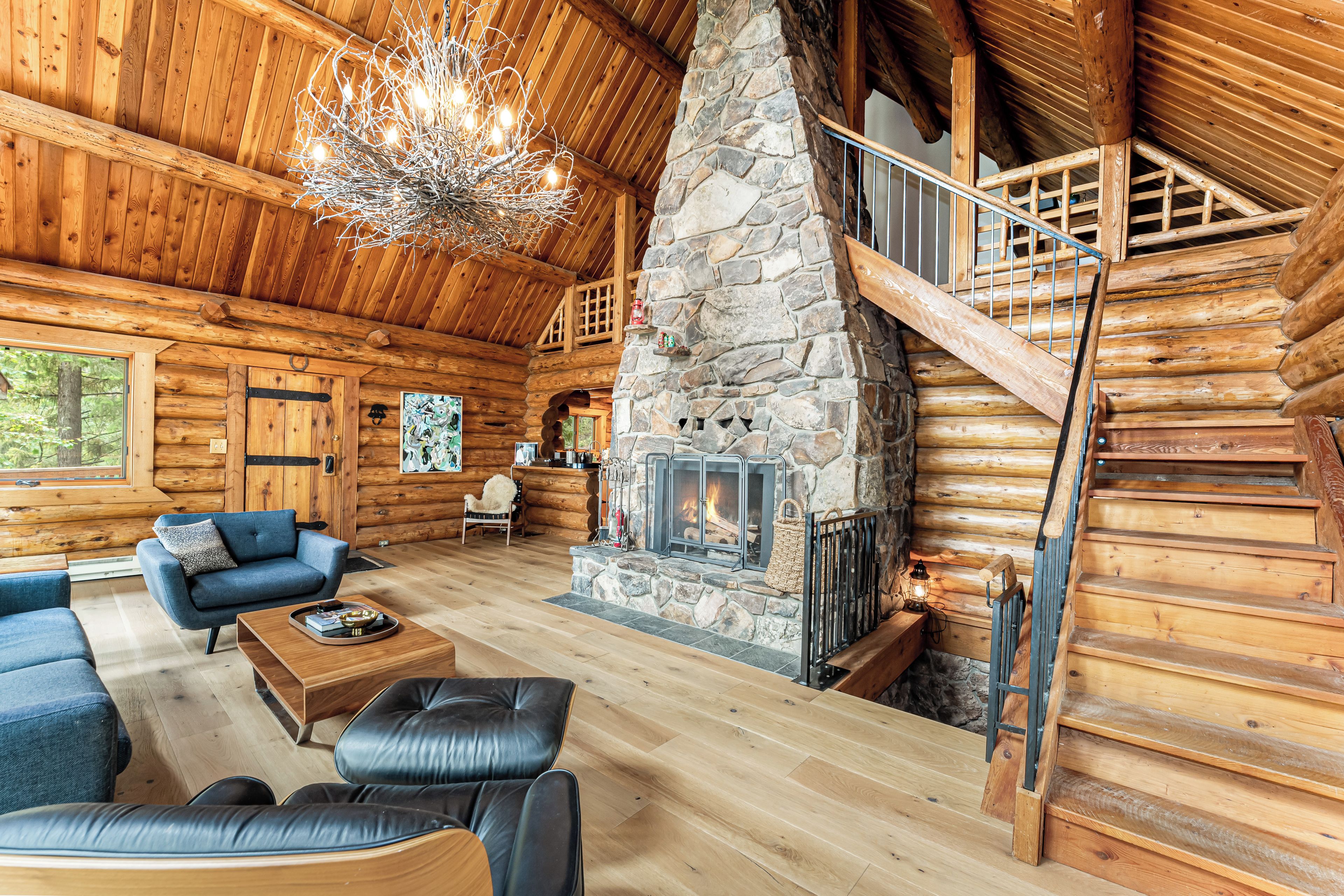 L’intérieur d’une cabane en rondins, avec une cheminée en pierre, un escalier en bois et un lustre.