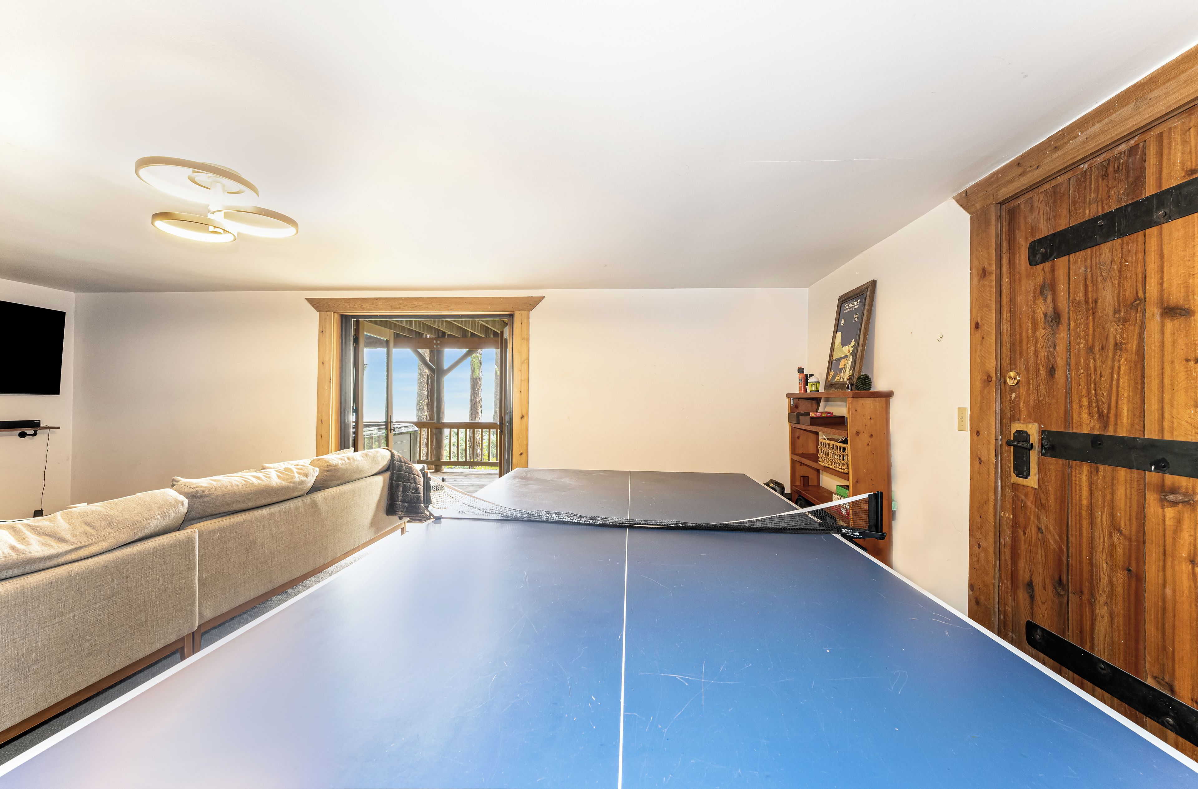 Une table de ping-pong se trouve dans une pièce qui contient un canapé, une porte en bois et une fenêtre donnant sur des arbres.