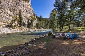 A river with clear water, surrounded by rocky cliffs and dense forest. There are outdoor seating areas with blue chairs and tables. - Riverfront Retreat | Hot Tub | Fire Pit | Large Groups | Fishing (Gallatin Gateway)