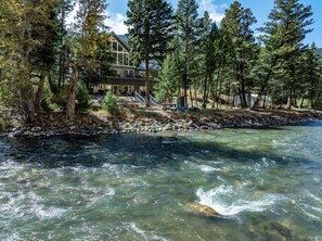 A riverside lodge with a stone retaining wall, a wooden deck, and a clear view of the river. - Riverfront Retreat | Hot Tub | Fire Pit | Large Groups | Fishing (Gallatin Gateway)