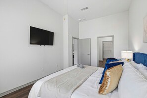 Ein Hotelzimmer mit Bett, Flachbildfernseher an der Wand, Nachttisch mit Lampe und einer Tür zu einem weiteren Raum.