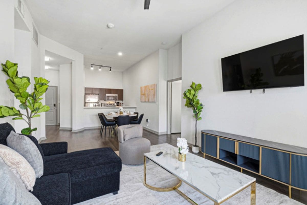 Un salon moderne avec un canapé, un pouf et une table basse. On y trouve également un coin repas avec une table et des chaises, et une cuisine en arrière-plan. Un téléviseur à écran plat est fixé au mur.