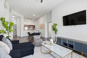 Un salon moderne avec un canapé, un pouf et une table basse. On y trouve également un coin repas avec une table et des chaises, et une cuisine en arrière-plan. Un téléviseur à écran plat est fixé au mur.