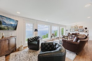 Ein großzügiges Wohnzimmer mit Panoramafenster und Blick auf die Berge, mit Ledersofas, einem Holzkasten und einem flachbildfernseher an der Wand.