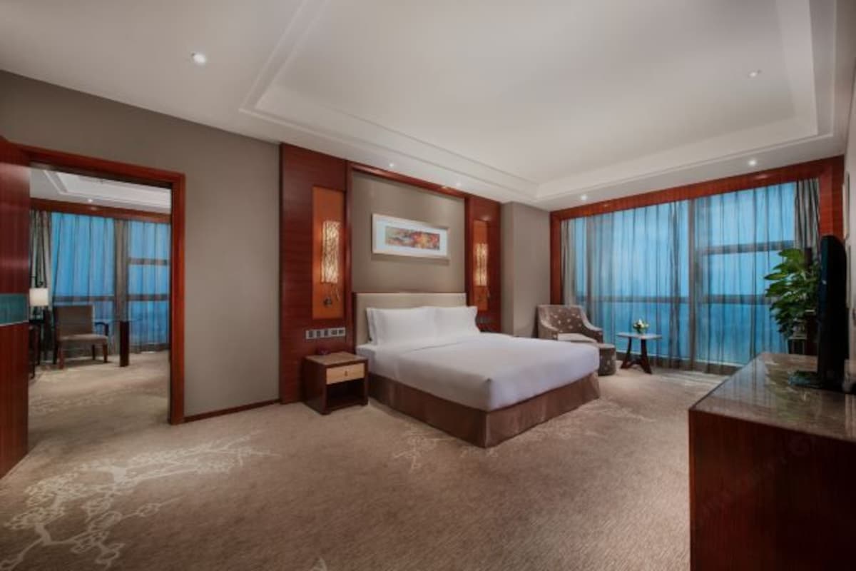 Una habitación de hotel con una cama grande, un escritorio, una silla, una mesita y un ventanal con cortinas.