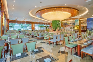 A spacious restaurant with multiple dining tables and chairs, a central decorative light fixture, and a green wall with plants.