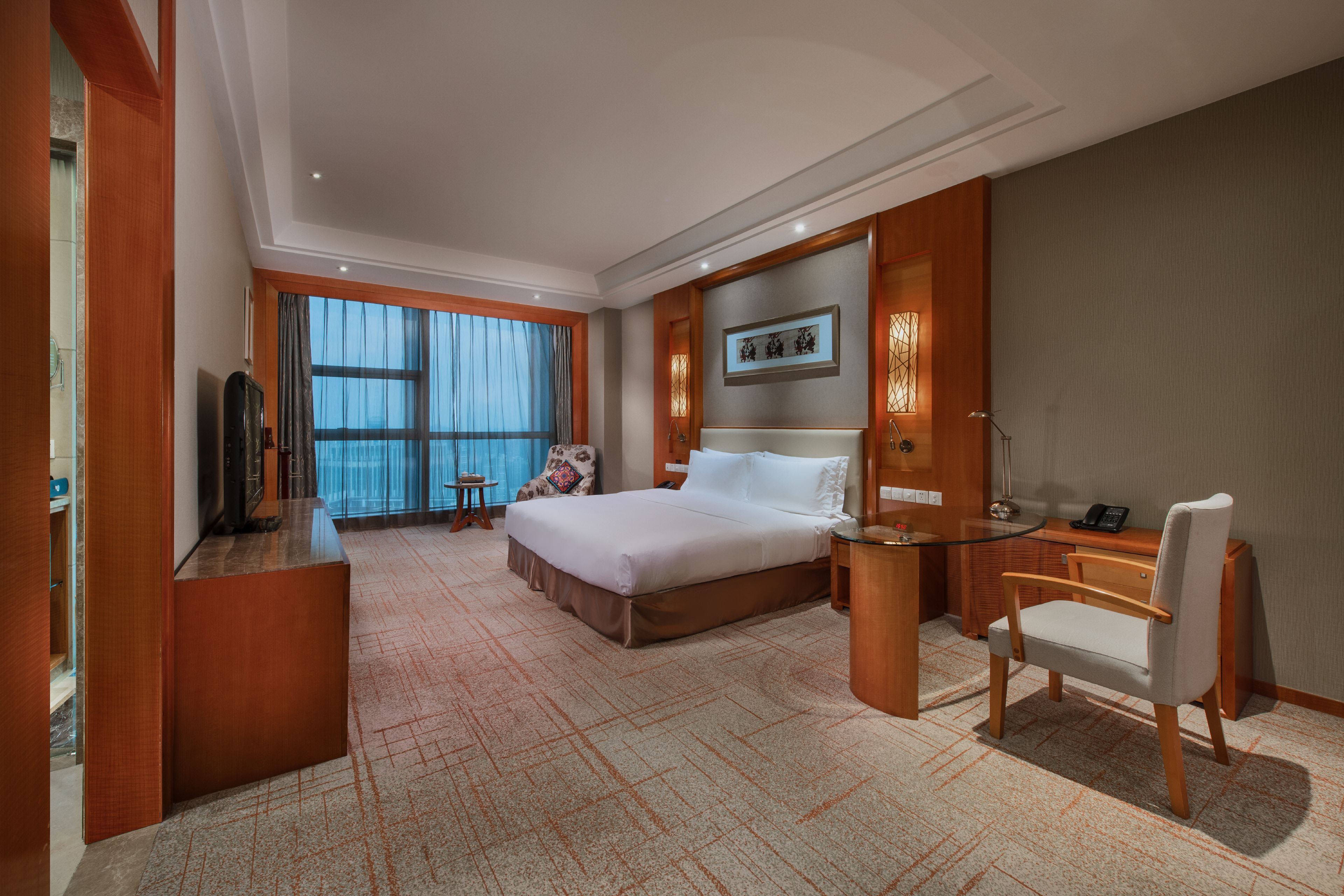 A hotel room with a large bed, a desk with a chair, a television, and a large window with curtains.