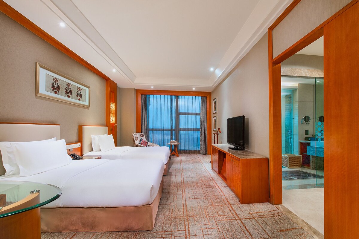A hotel room with a large bed, a TV, a desk, and a view of the sea through the window.