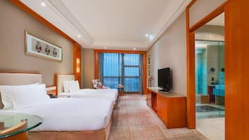 A hotel room with a large bed, a TV, a desk, and a view of the sea through the window.