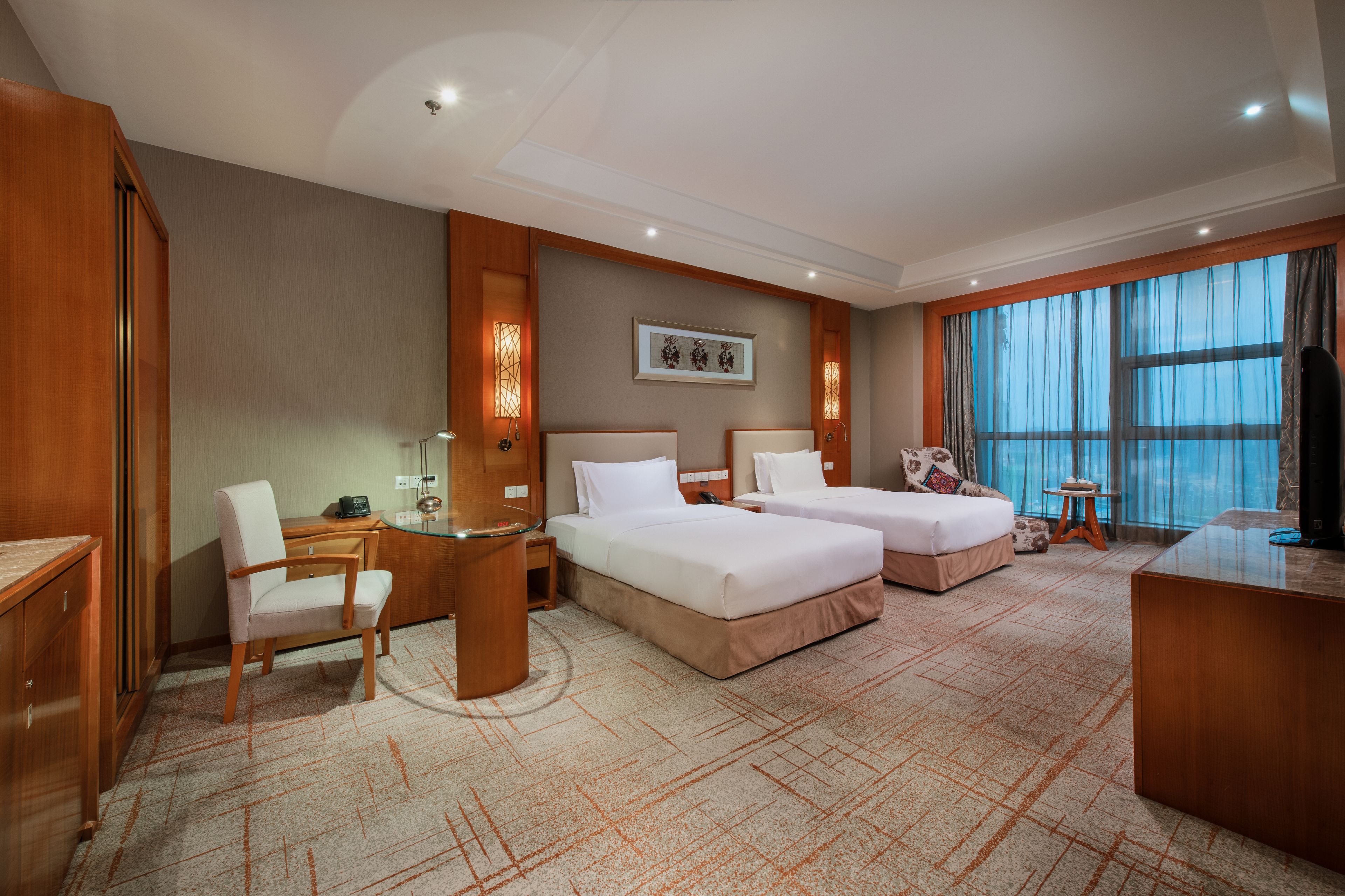 A hotel room with two beds, a desk, a chair, a television, and a large window with curtains.