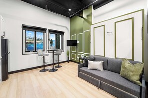 A modern living room with a grey sofa, two bar stools, a flat-screen TV, and a large window with a view of the ocean. - Experience Downtown Energy Near Petco Park & Dine! (San Diego)
