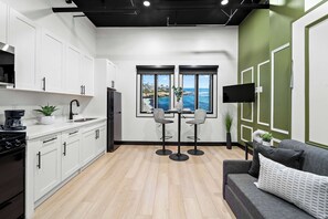 A modern kitchen with white cabinets, a black refrigerator, and a coffee maker. There are two bar stools and a dining table with a view of the ocean through large windows. - Experience Downtown Energy Near Petco Park & Dine! (San Diego)