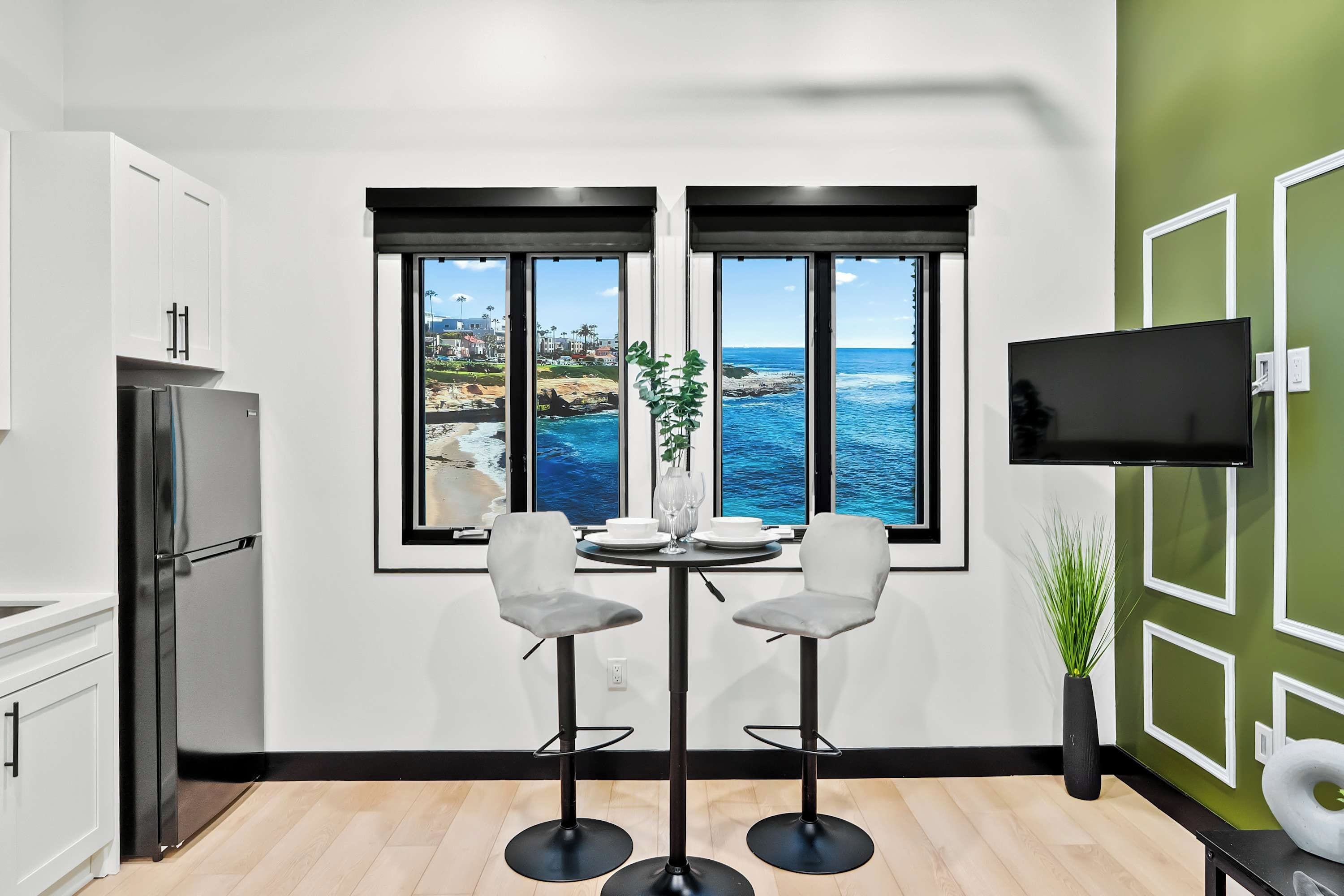 A modern kitchen with a large window overlooking the ocean, a dining area with two chairs, and a flat-screen TV mounted on the wall.
