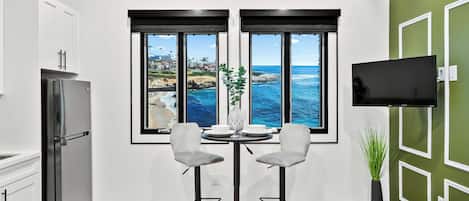A modern kitchen with a large window overlooking the ocean, a dining area with two chairs, and a flat-screen TV mounted on the wall.