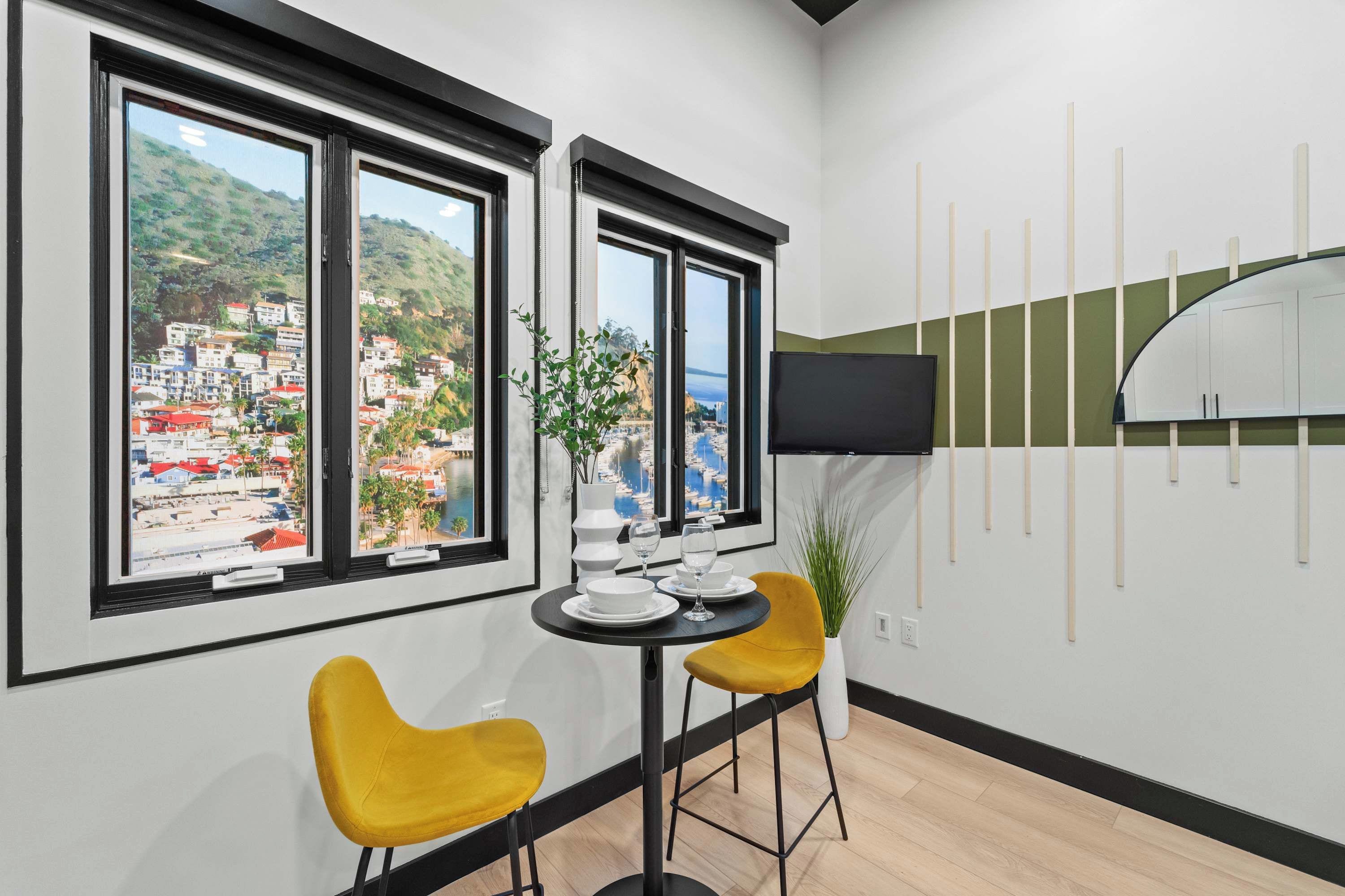 A modern room with two large windows, a flat-screen TV mounted on the wall, and a small dining table with yellow chairs.