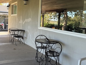 Un área de estar al aire libre con sillas de metal y una mesa, una ventana con vistas a follaje, y un vehículo estacionado al fondo.