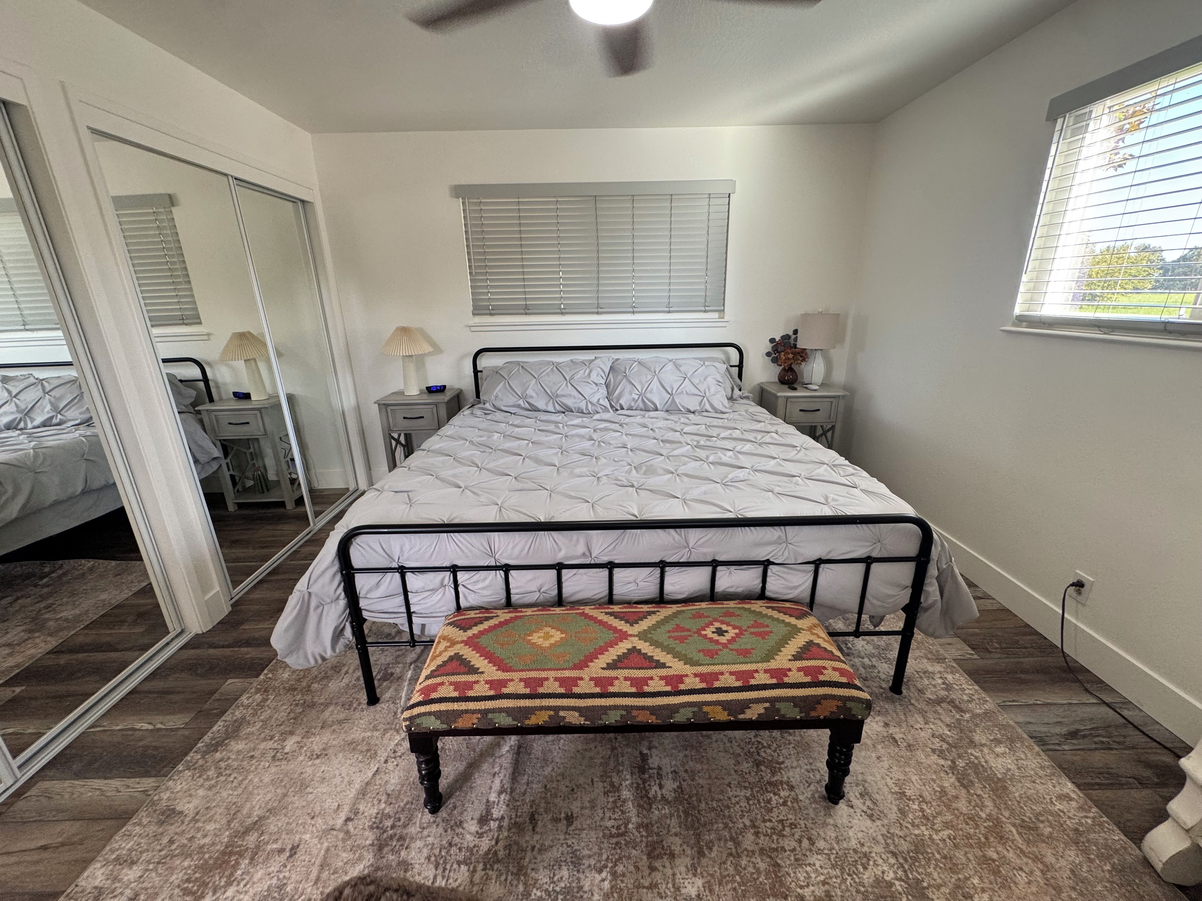 Une chambre à coucher comprenant un lit, des tables de chevet, un banc, un ventilateur de plafond et une fenêtre avec des stores.