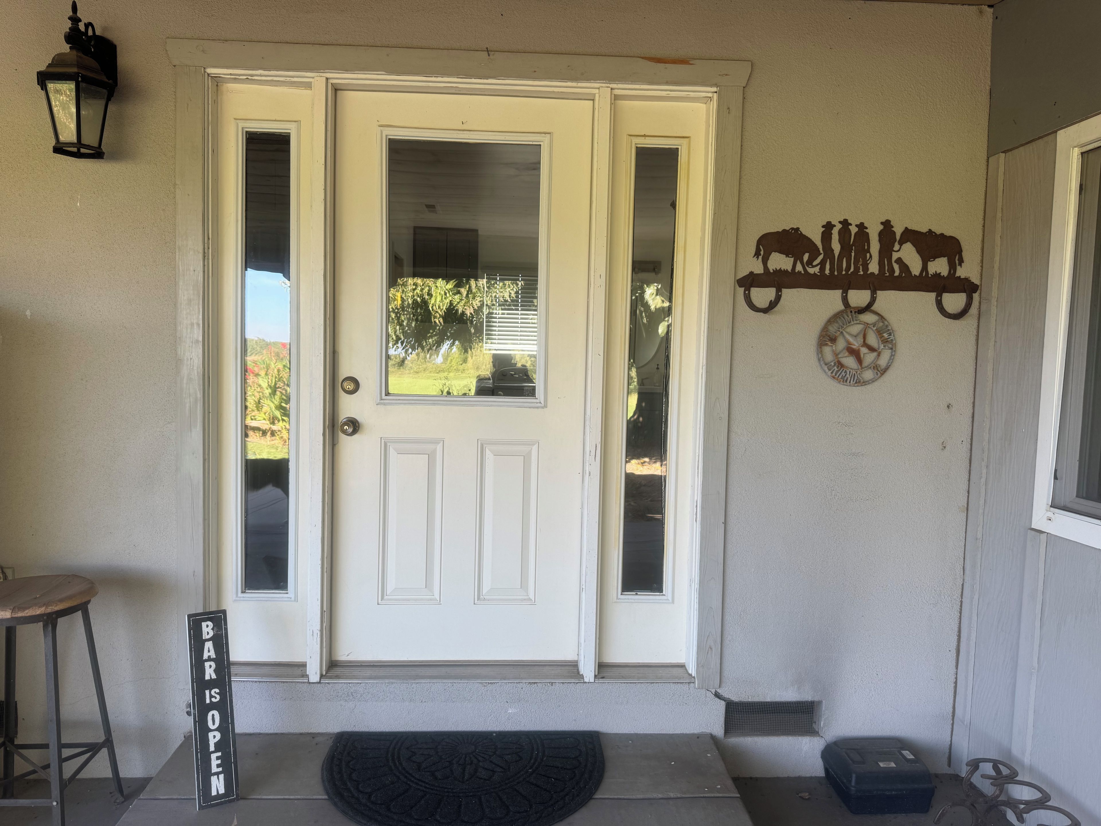 A white front door with a glass panel, a black bar sign, a metal horse decoration, and a small stool.