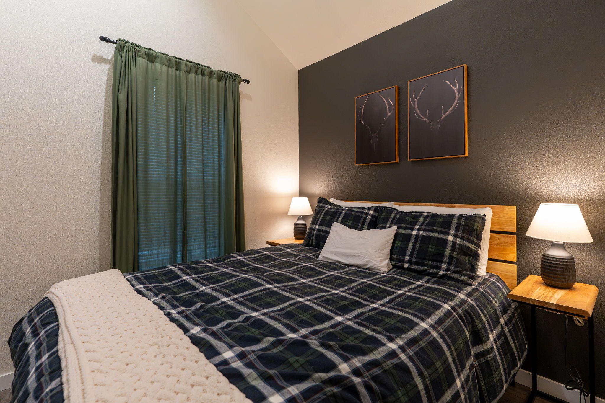 A bedroom with a bed, two wall-mounted lamps, a window with curtains, and two framed pictures on the wall.
