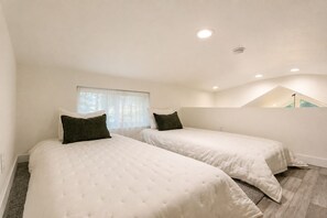 Ein Schlafzimmer mit zwei Betten, einem Fenster mit durchsichtigen Vorhängen und einer schrägen Decke mit Einbaustrahlern.