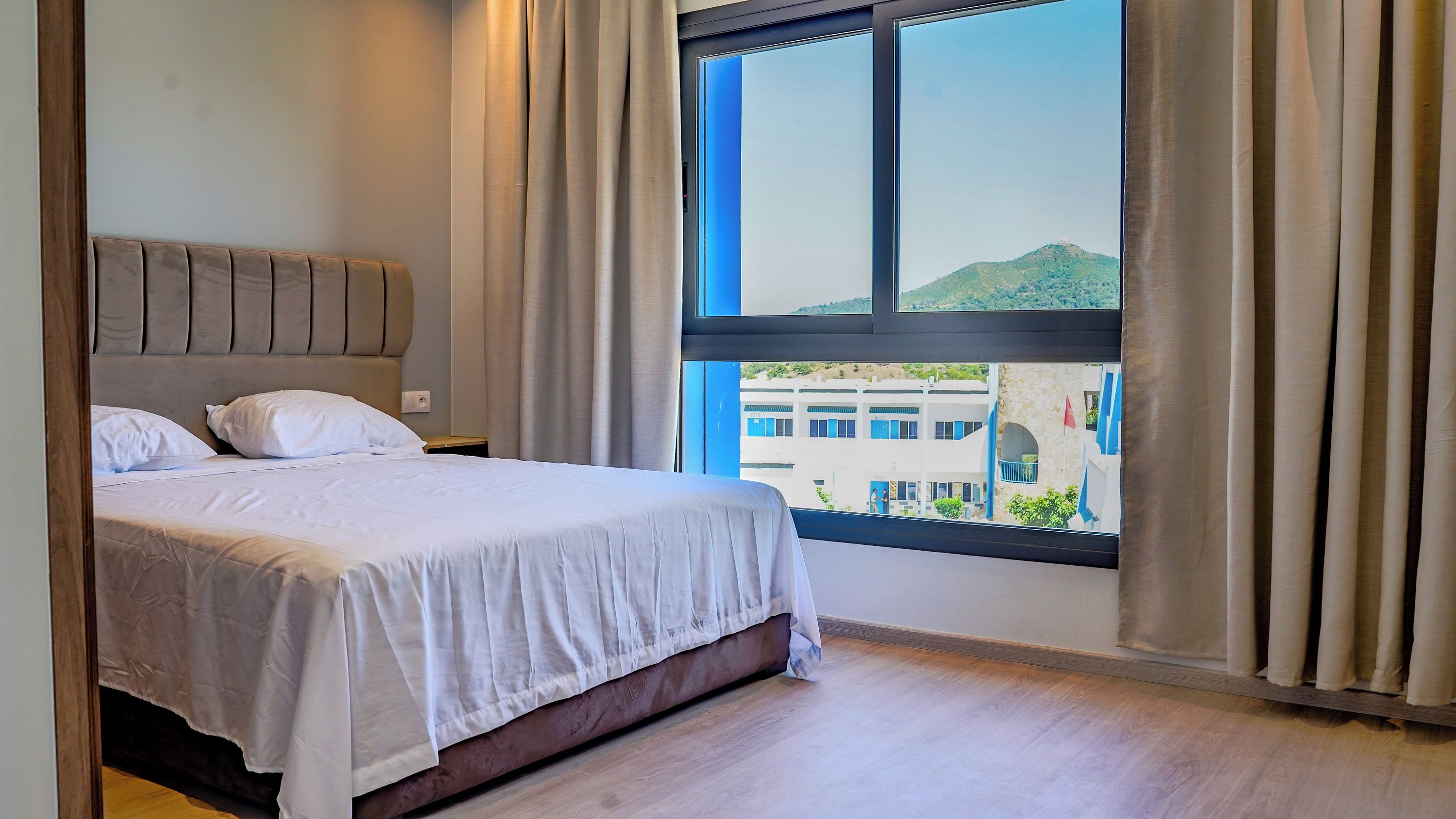 Una habitación de hotel con una cama, un ventanal con cortinas y vistas a edificios.