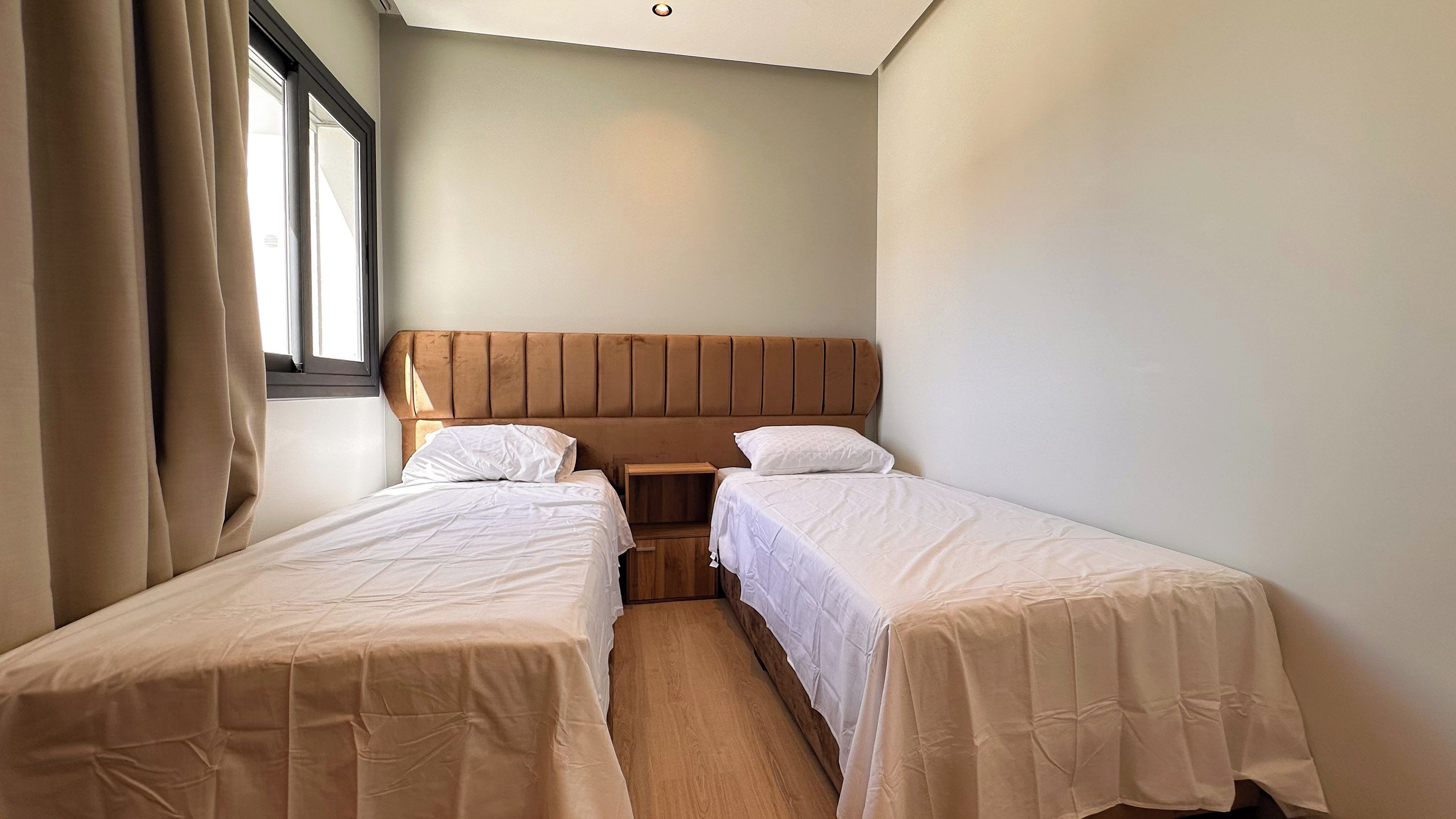 Ein Doppelzimmer mit zwei Einzelbetten, einem Holzkopfstück, einem Nachttisch und einem Fenster mit Vorhängen.