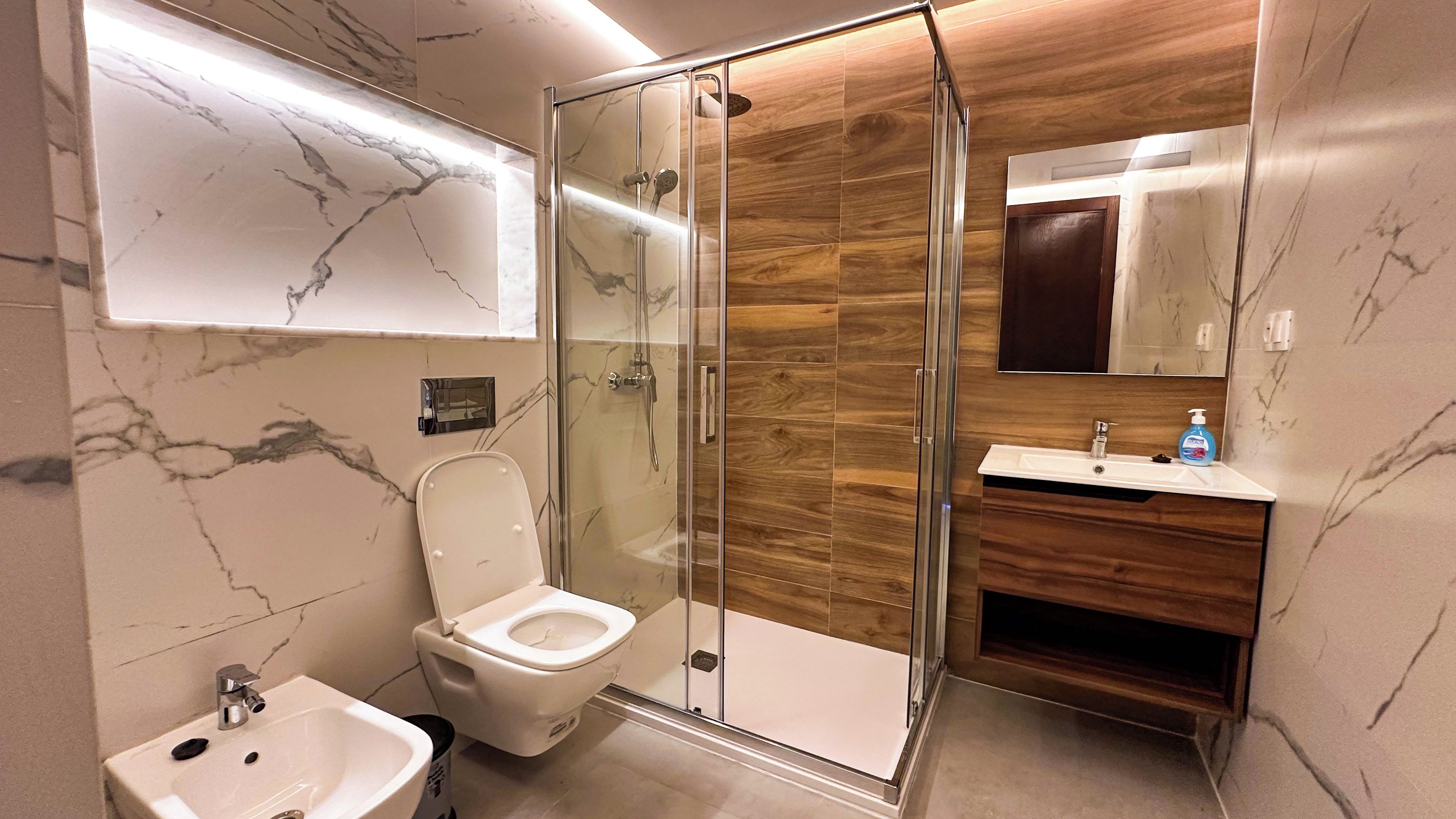A modern bathroom with a glass-enclosed shower, a marble-tiled wall, a wooden vanity, and a white sink.