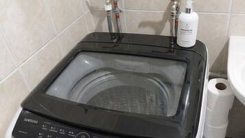 A Samsung washing machine with a transparent door, control panel, and two red valves, located in a bathroom with a tiled wall and a white sink.