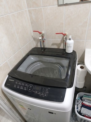 Una lavadora Samsung con puerta transparente, panel de control y dos válvulas rojas, ubicada en un baño con pared de azulejos y un lavamanos blanco.
