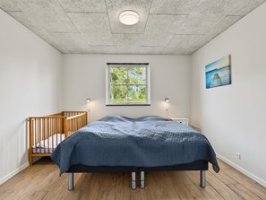 A bedroom with a wooden crib, a bed with a blue quilt, a window with a view of greenery, and a wall-mounted painting. - "Nina" - 600m from the sea by Interhome (Væggerløse)