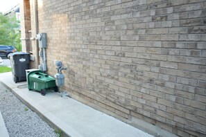 A building exterior with a brick wall, a green dumpster, a black trash bin, and a utility meter. - Cozy Basement Suite in Brampton | Modern & Central (Brampton)