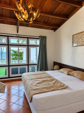 A hotel room with a large bed, a wooden ceiling, a view of the sea through multiple windows, and a decorative wall piece. - Rancho frente al mar - Piscina + Pádel - BIG KAHUNA SURF CITY (La Libertad Costa)