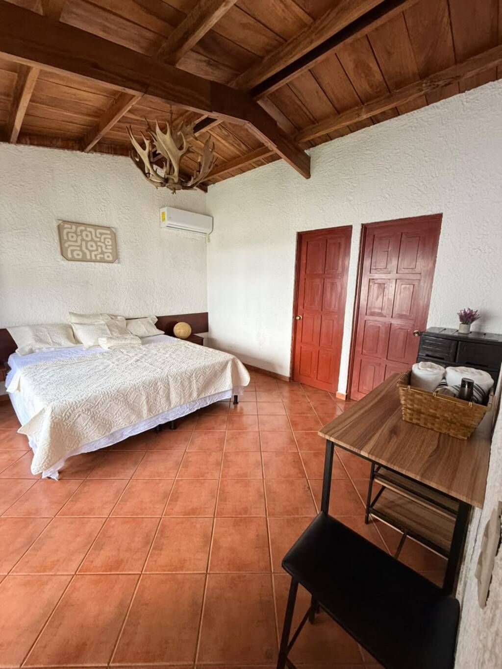 A bedroom with a bed, a wooden table, a chair, a wardrobe, and a ceiling with wooden beams.