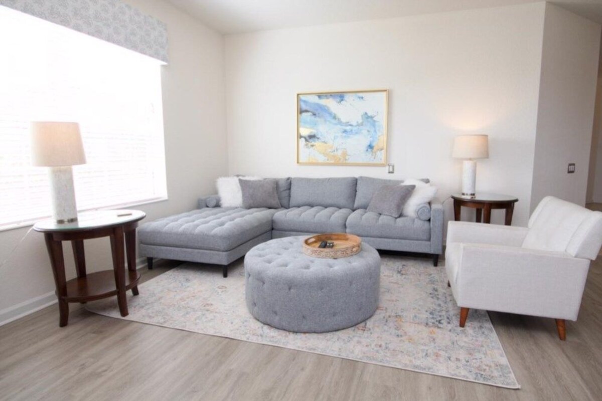 A living room with a grey sectional sofa, a round ottoman, a white armchair, and two side tables with lamps.