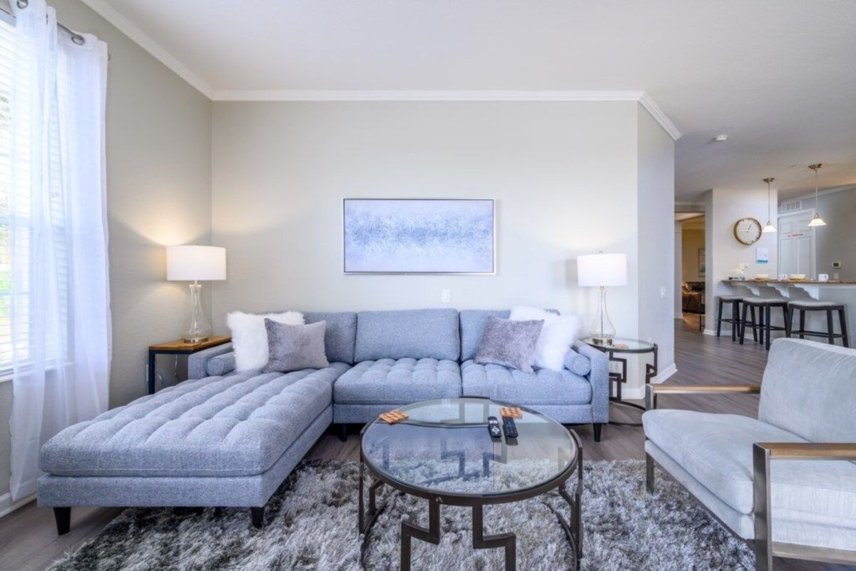 A modern living room with a sectional sofa, a glass coffee table, and a dining area in the background.