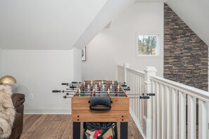 A room with a foosball table, a sofa, a lamp, and a staircase with a stone wall. - Creek Cabin, Hot Tub + Fire Pit + Private Deck + Games (Ellijay)