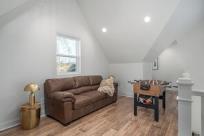 A cozy room with a brown leather sofa, a foosball table, and a window with a view of trees. - Creek Cabin, Hot Tub + Fire Pit + Private Deck + Games (Ellijay)