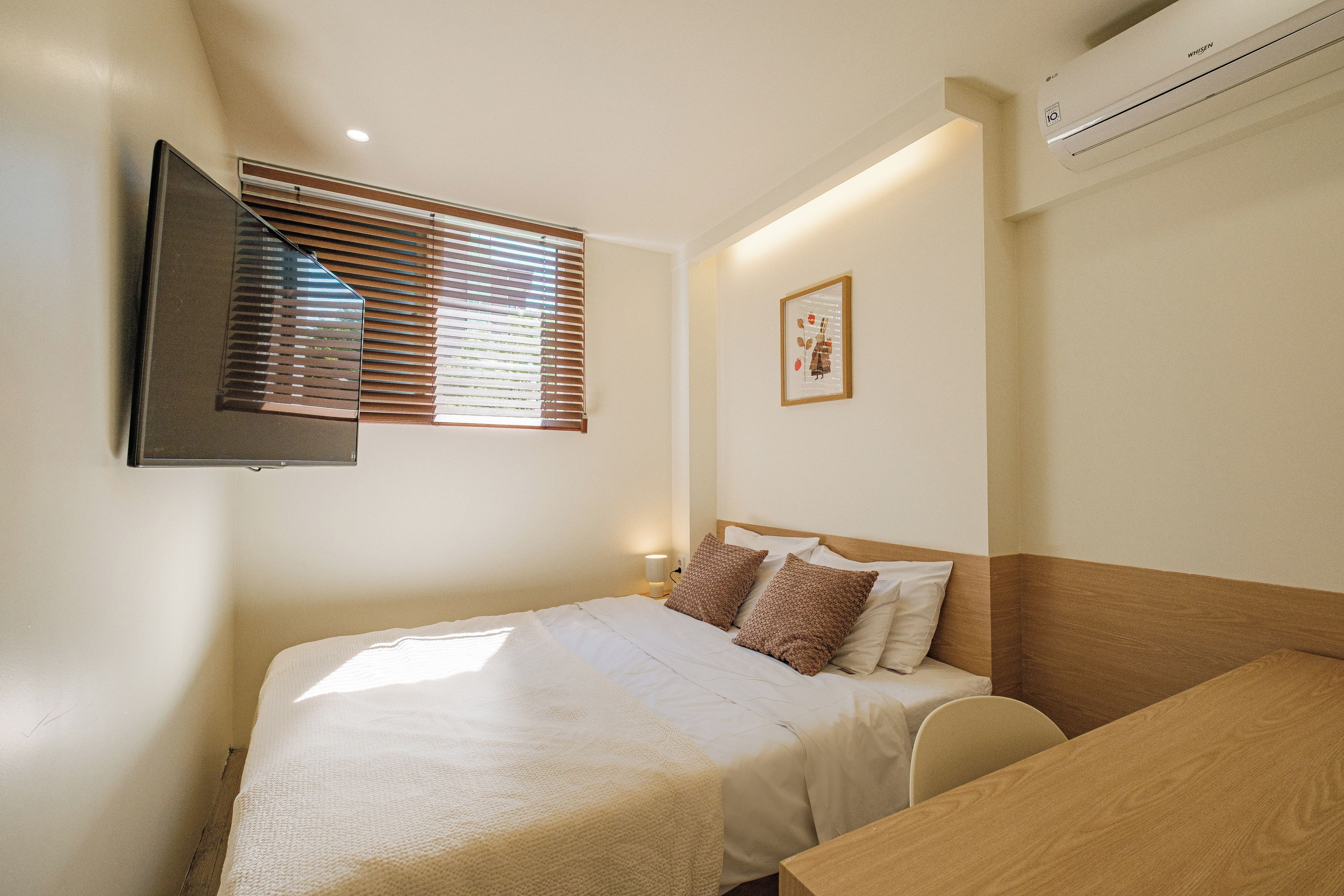 A hotel room with a bed, a TV mounted on the wall, a window with blinds, a framed picture on the wall, and an air conditioning unit.