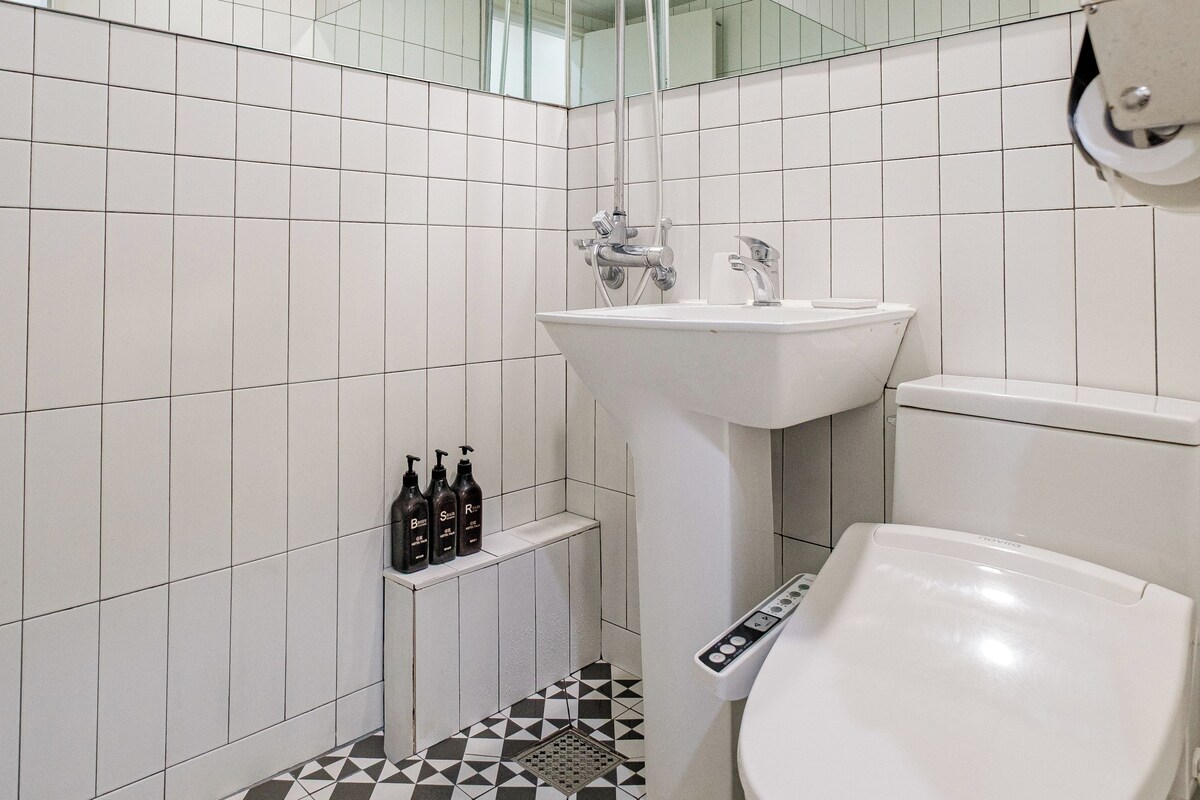Uma casa de banho moderna com parede revestida a azulejos brancos, um espelho grande, chuveiro, pia e sanita.