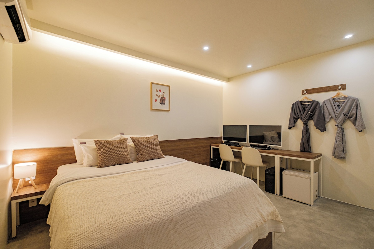 Quarto de hotel com cama, escrivaninha, computador e televisão fixada na parede.