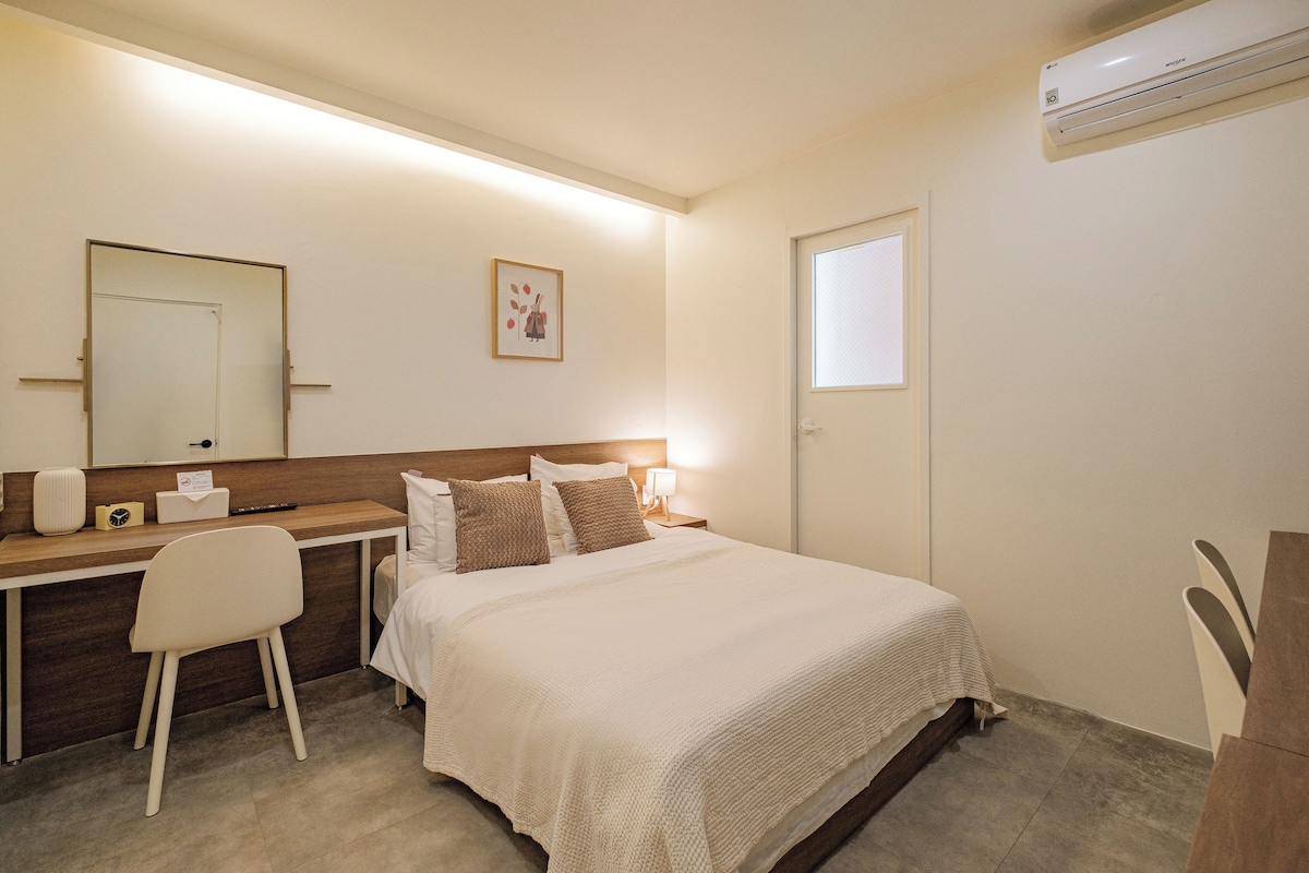 Quarto de hotel com cama, mesa de cabeceira, cadeira, escrivaninha e ar condicionado fixado na parede.