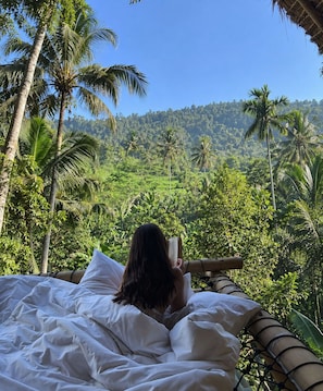 A person lying on a bed with a view of a lush green landscape and palm trees. - Kayun Sidemen (Sidemen)