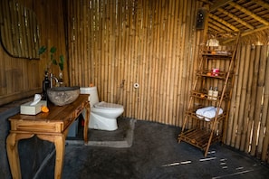 A bamboo-walled room with a wooden table, a stone basin, a toilet, and a shelf with toiletries. - Kayun Sidemen (Sidemen)