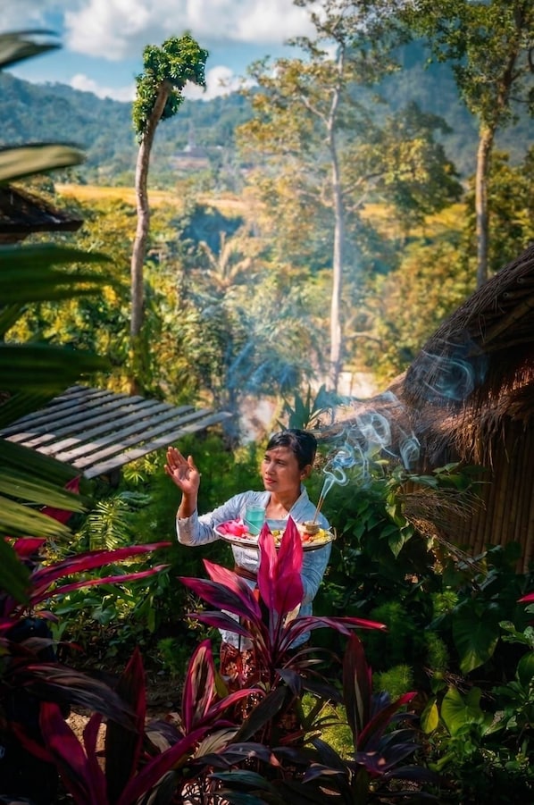 A person in a garden with a tray of food and drink, surrounded by lush greenery and a bamboo structure.