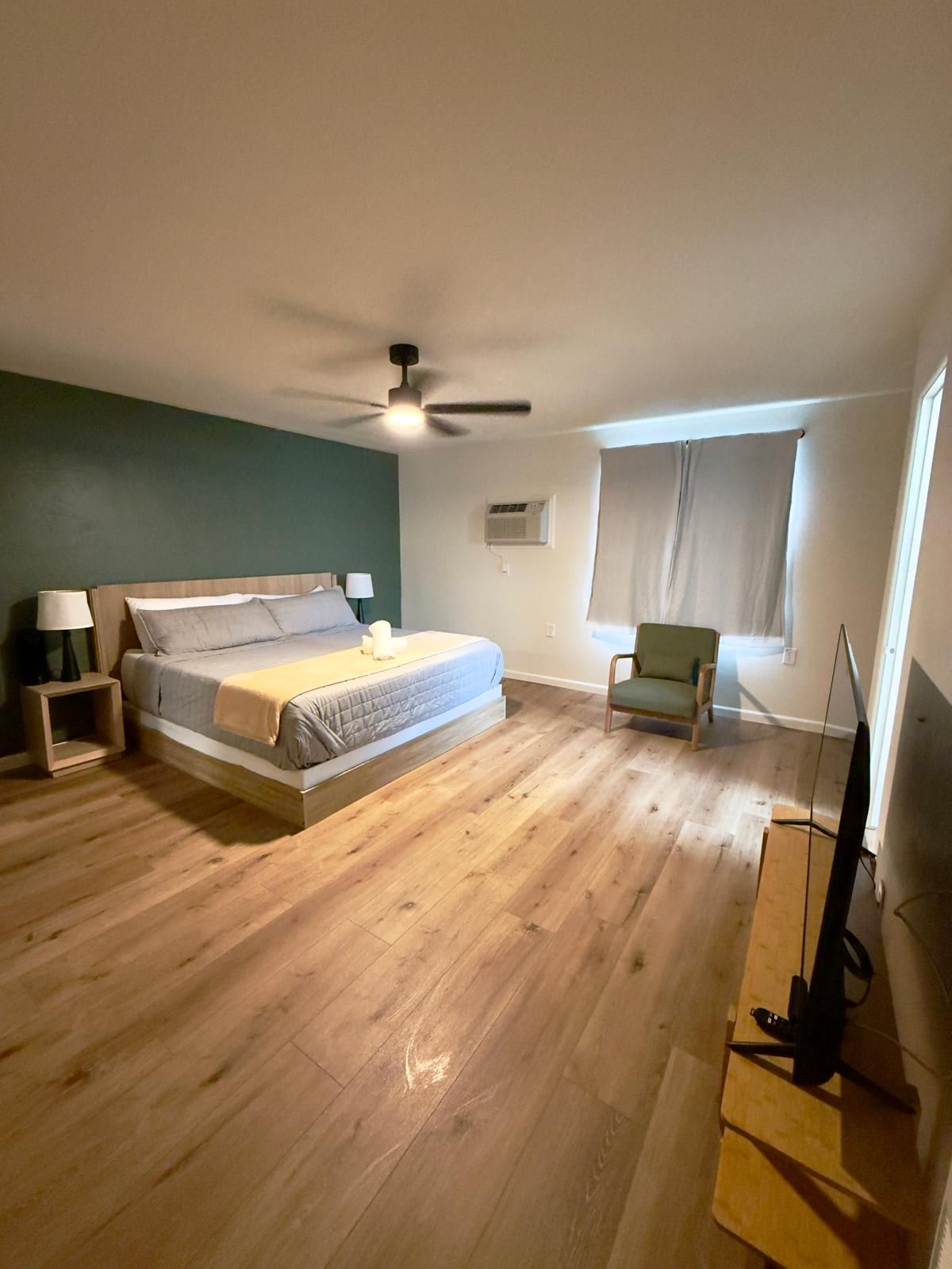 A bedroom with a bed, a ceiling fan, a chair, a window, and a TV stand.