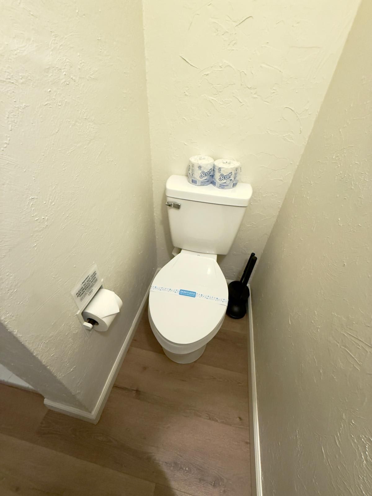 A small bathroom with a toilet, two rolls of toilet paper, a toilet brush, and a paper towel holder.